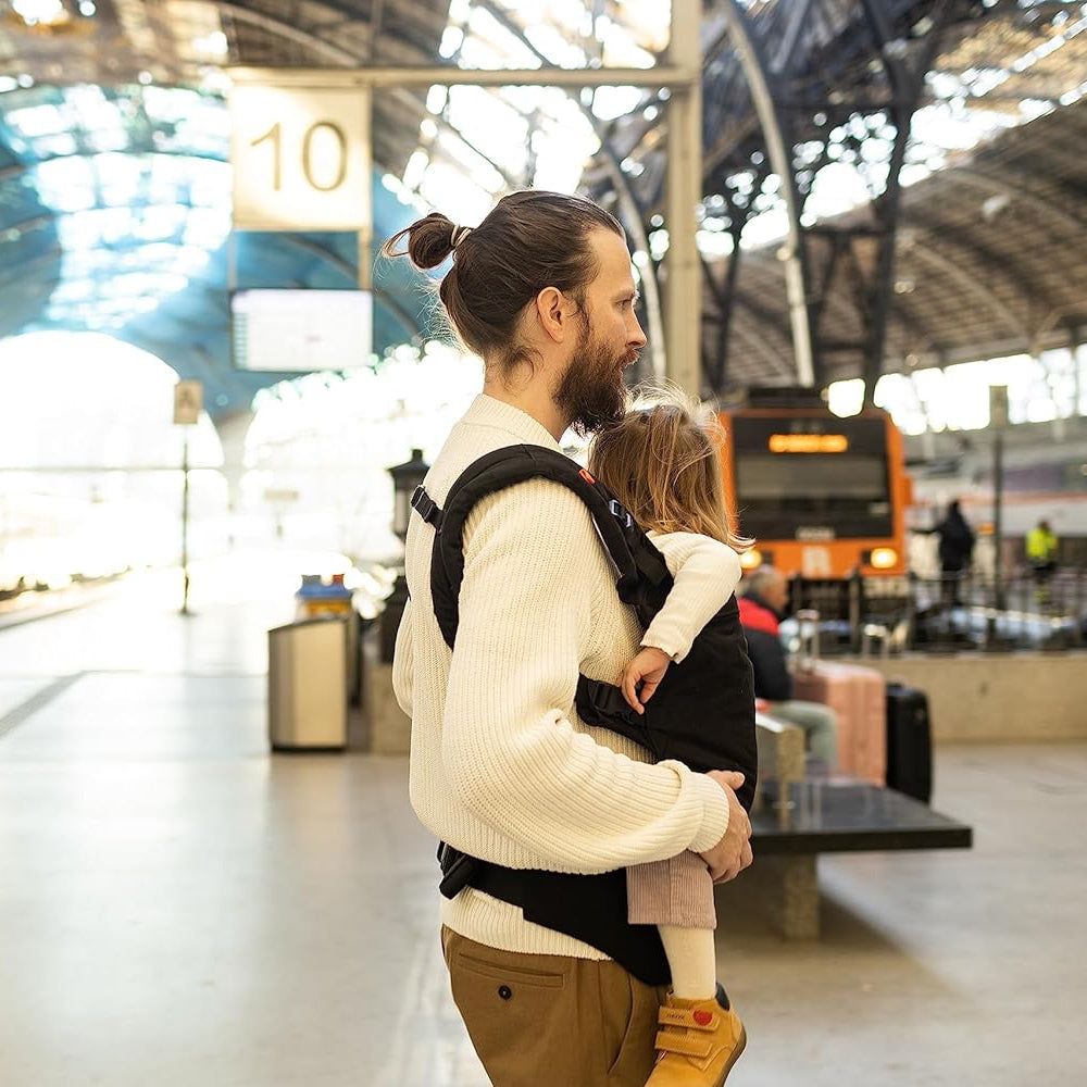 Man carrying a child in a toddler carrier at a train station.