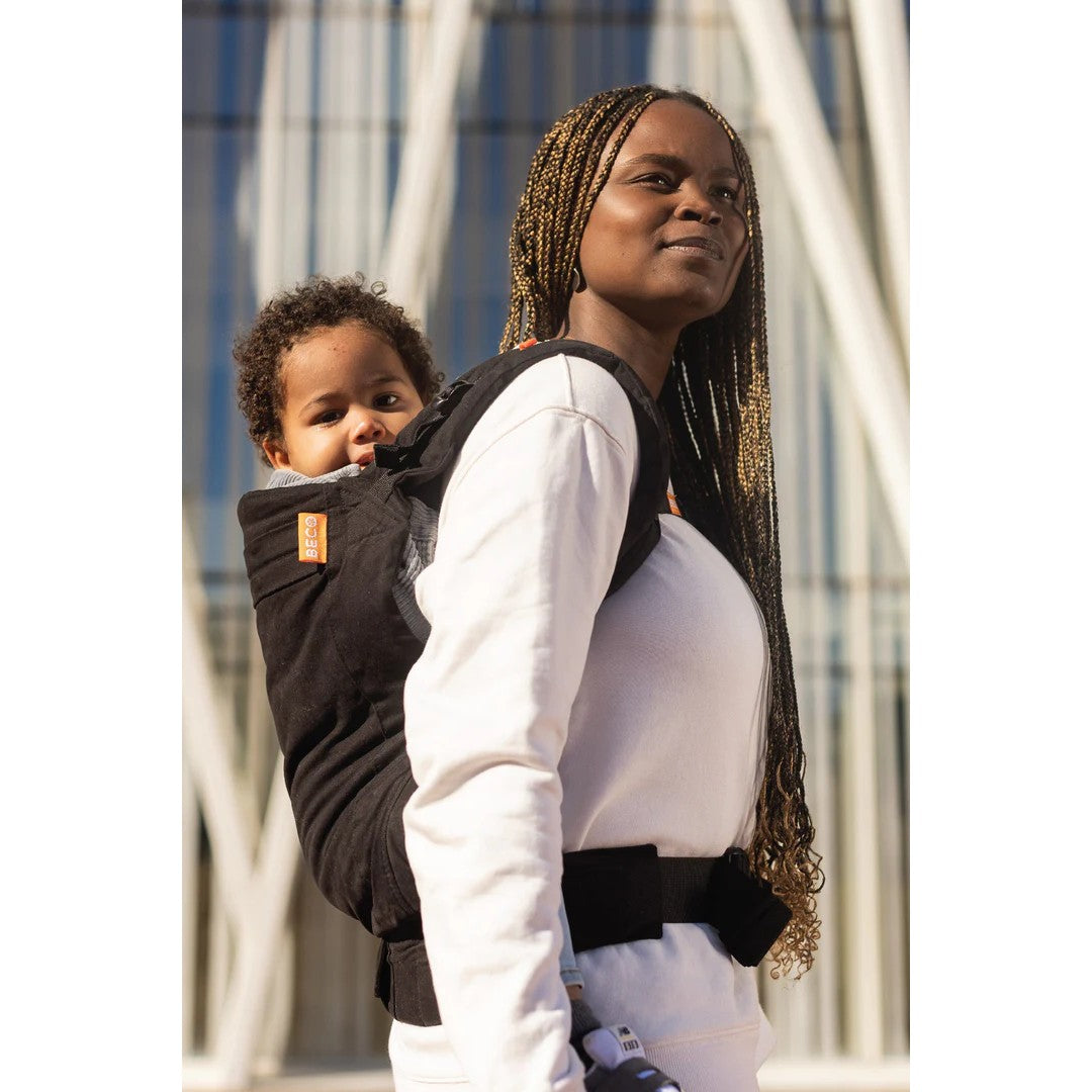A woman wearing a black baby carrier with a child on her back, photographed outdoors in front of a modern glass building.